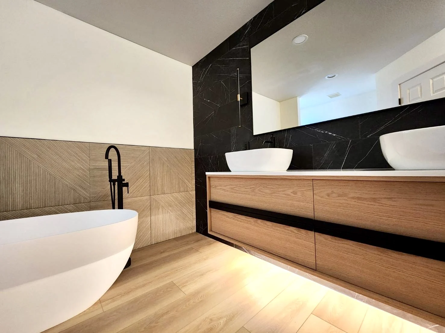 Bathroom after renovation: floating white-oak double vanity with vessel sinks, black marble feature wall, freestanding tub and wood-look floor